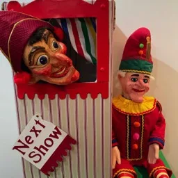 Fotografía de una caseta de Punch & Judy con la gran cabeza de Mr Punch asomando. Un cartel 'Próximo Espectáculo' está clavado en la caseta.