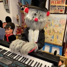 Un autómata peludo con sombrero de copa tocando un teclado, rodeado de marionetas y recuerdos.
