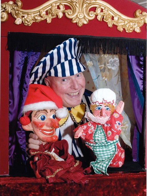 Martin Scott Price in its Puch & Judy booth holding Mr Punch & Judy puppets.